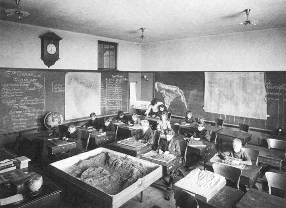 An elementary geography class at John Dewey's Laboratory School: children at individual desks in rows, each desk holding a tray of sand and clay, a large relief model on the floor, blackboards and maps covering the walls, and an adult standing among the children at the back of the room leaning over a pupil.